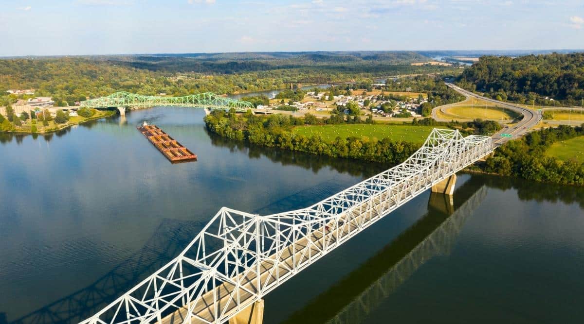 A river Tugboat pushes barge contents down the Ohio River. View from above of the wide Ohio River with two long white and green bridges leading to the small town of Gallipolis in Ohio.