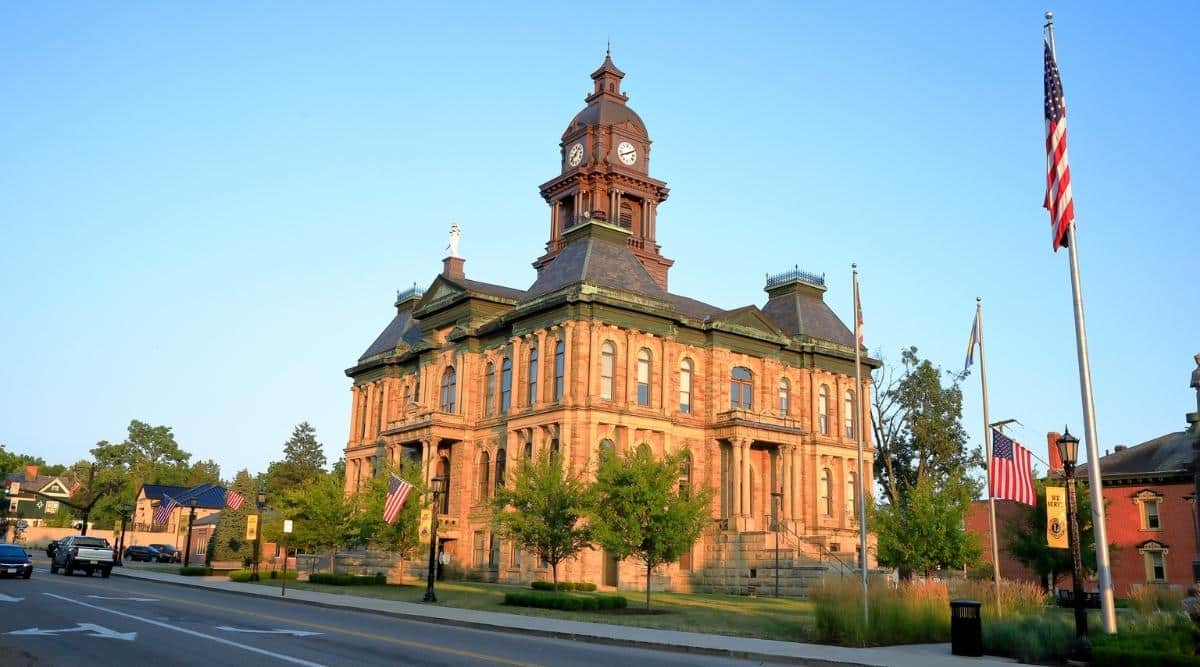 The Holmes County Courthouse in Millersburg, Ohio was built in the Victorian Gothic architectural style, featuring a red brick exterior, a clock tower with a four-faced clock, and intricate stonework details such as arched windows and ornate cornices. There are many American flags near the building.