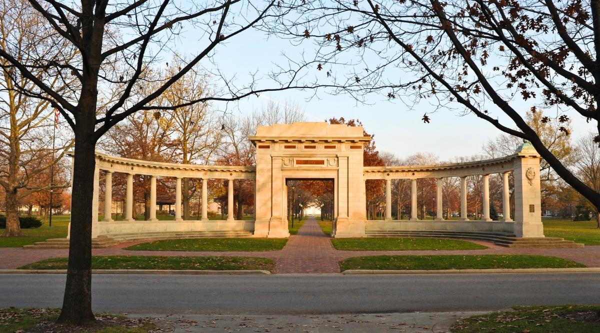 The Memorial Arch in Tappan Square, Oberlin College is a historic landmark, featuring a neoclassical architectural style with a red brick exterior, a large central arch flanked by columns, and a bronze plaque commemorating the names of the fallen soldiers. Around the arch there are beautiful manicured green lawns and trees with orange leaves.