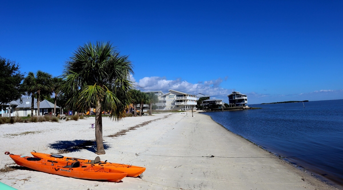 Cedar Key is a small, picturesque island located on the west coast of Florida, known for its natural beauty, laid-back atmosphere, and unique history. The beach has white sand, blue water, tropical palm trees and various white buildings with shops, hotels and entertainment. Two bright orange kayaks stand on the shore.