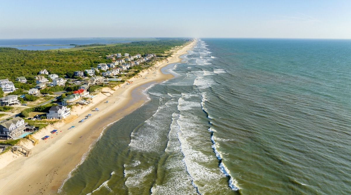 Corolla Beach in North Carolina is a beautiful, secluded beach located on the Outer Banks, known for its wide expanse of sandy beaches, crystal-clear waters, and wild horses that roam the area. Top view of the rippling ocean, golden sandy beach and small cottages.