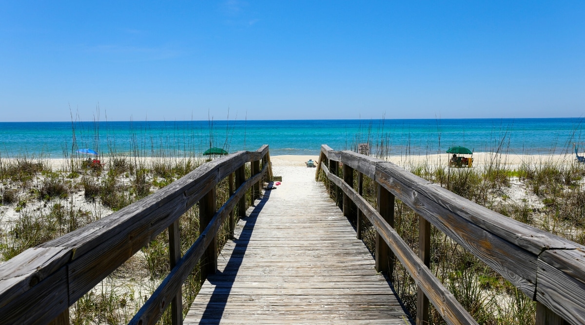 Henderson Beach State Park in Florida is a beautiful natural area with white sandy beaches, crystal-clear waters, and nature trails for hiking and wildlife viewing. Access to the beach via a wooden bridge, after which a white beach and clear blue waters open.