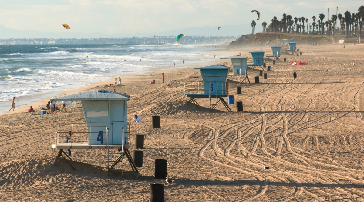 Huntington State Beach in California is a popular spot for surfing and swimming, with its wide sandy shoreline and consistent waves. Close-up of a beach with golden sand, big blue and white waves and beach lifeguard towers.