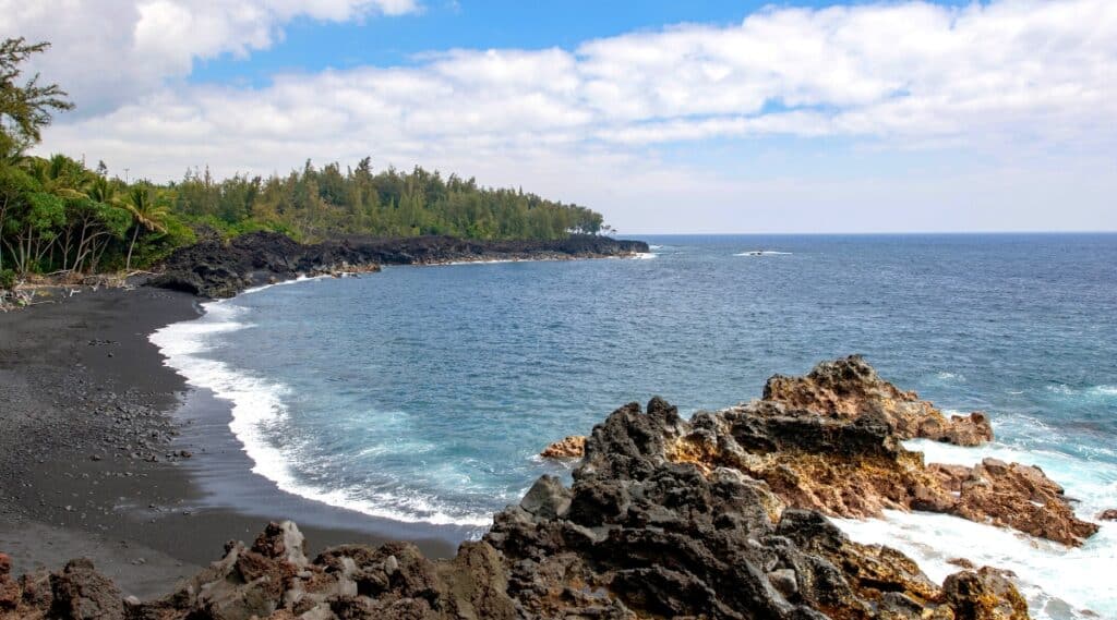 Kehena Beach is a secluded and beautiful black sand beach located on the Puna coast of Hawaii's Big Island. The water is crystal clear, blue. The beach is surrounded by tropical vegetation.