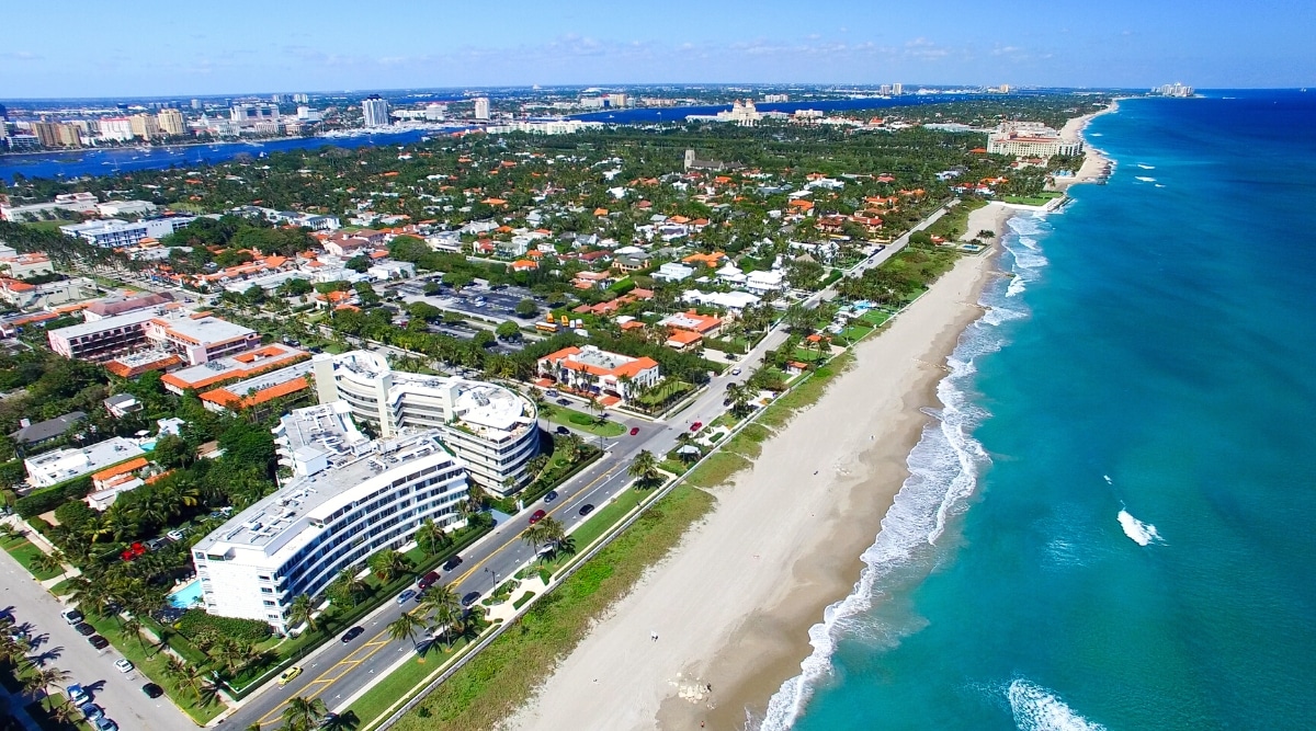 This image presents a panoramic view of the exquisite Palm Beach, Florida. The picture offers a bird's-eye view of the sandy coastline that extends alongside the crystal-clear waters of the Atlantic Ocean, with the city in the background adorned with lush vegetation. The beach is flanked by a lush, meticulously landscaped promenade with palm trees.