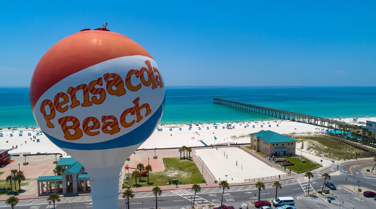 Pensacola Beach is located on Santa Rosa Island, with pristine white sand beaches and crystal clear turquoise water. In the foreground, one of the beach's most iconic landmarks is the Pensacola Beach Water Tower, a towering white structure that serves as a beacon for visitors. The beach is also known for its lively atmosphere, beachfront restaurants, bars, shops, long pier, and various outdoor activities such as kayaking, kayaking, and fishing.