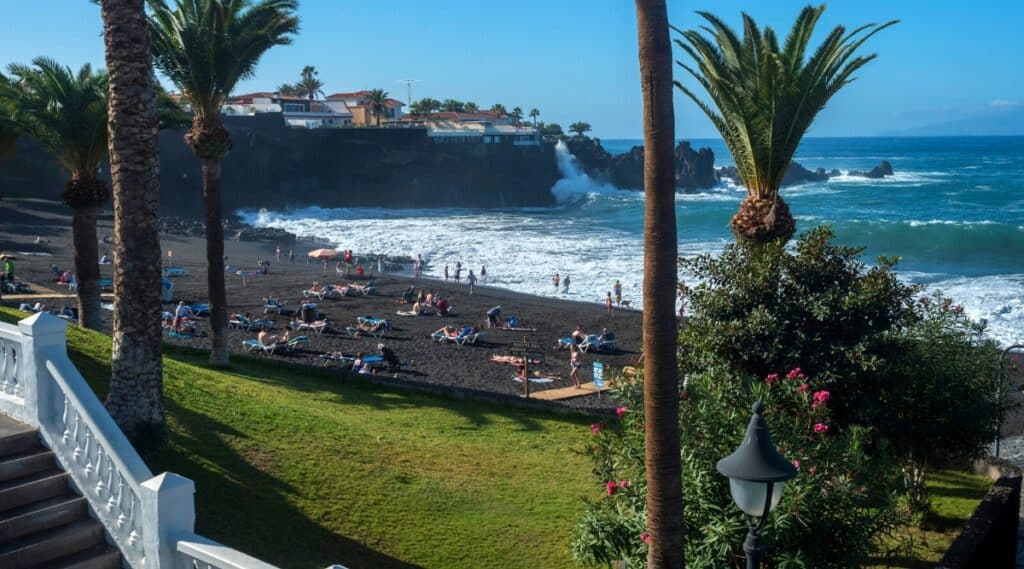 Playa de la Arena is a black sand beach located on the west coast of Tenerife, one of Spain's Canary Islands. The beach is located in a sheltered bay and is known for its crystal clear waters and spectacular scenery with high cliffs and lush greenery. The beach is equipped with sun loungers, umbrellas, showers and wooden paths. Tall and beautiful palm trees and other flowering plants also grow on the beach.
