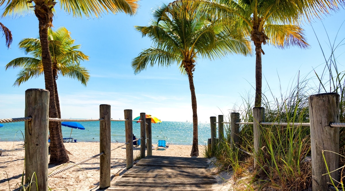 Smathers Beach is a popular beach located on the south shore of Key West in Florida. The beach offers a mile long stretch of soft sand and turquoise waters perfect for swimming and sunbathing. Tall tropical palm trees grow on the beach. Also, people relax with multi-colored sun umbrellas and sun loungers.