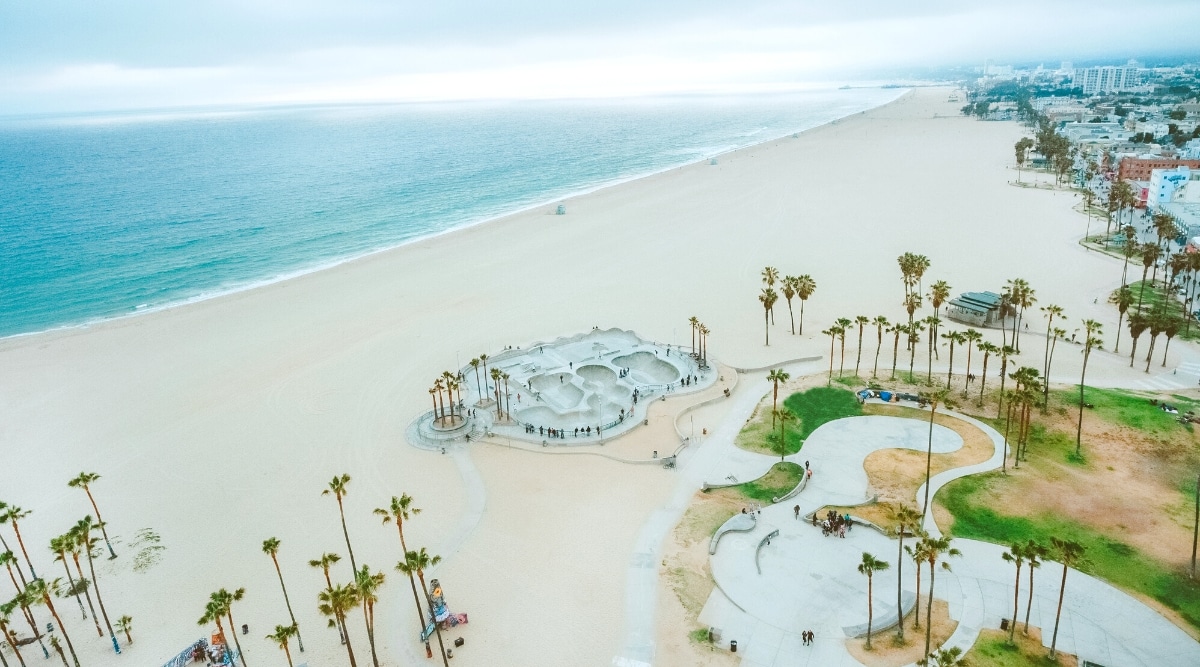 Venice Beach is a famous public beach located in the city of Los Angeles, California. Aerial view of wide sandy beach, blue clear waters, skate park, volleyball courts and fitness center.
