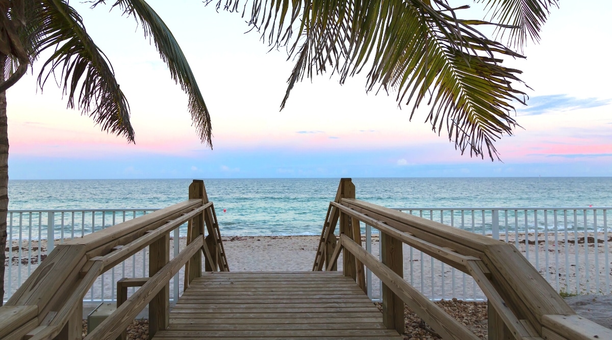 Vero Beach is a charming city located on the east coast of Florida. Close-up of the entrance to the beach at sunset with wooden steps and a beautiful fenced promenade with a white fence. The sand is white, the water is crystal blue, and the sky is iridescent shades of blue, white, and pink.