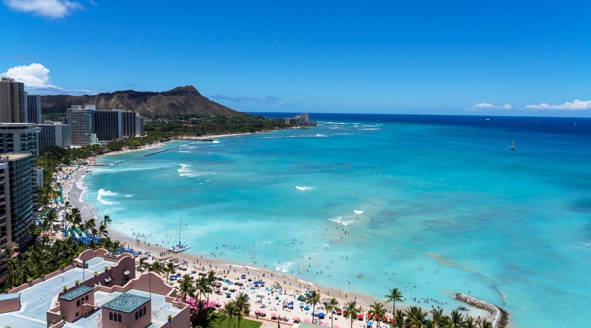 Waikīkī Beach in Oahu, Hawaii is a world-famous and popular destination known for its long sandy beach, and crystal-clear waters. Aerial view of a beach with crystal clear blue waters and white sands surrounded by tall hotels.