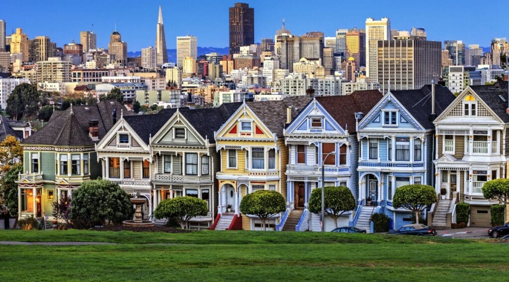Alamo Square Park in California with different colored houses and grass. The houses are green, yellow, blue, and white with a city view in the background.