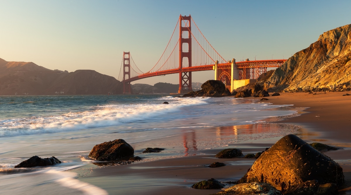 Golden gate bridge at sunset. Mountains in the background on the right hand side. Sun is hitting the bridge and mountains. Beach shores underneath the bridge are cascading against the beach at sunrise.