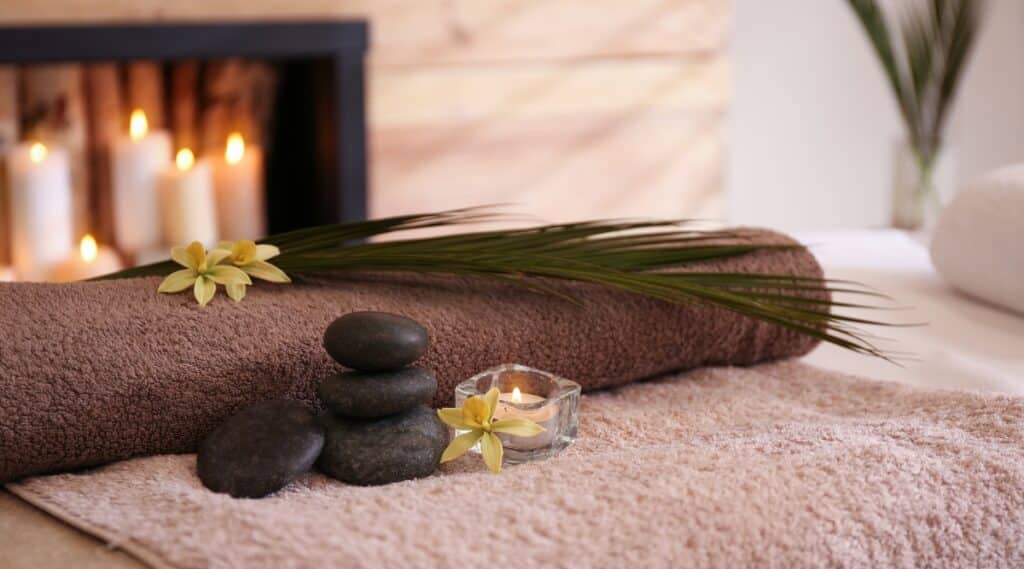 Folded massage towels with massage stones stacked next to them, topped with some palms and burning candles.