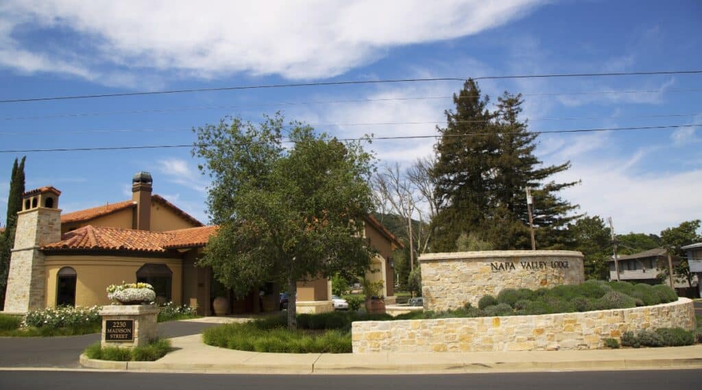 Street view of the Napa Valley Lodge in Yountbville, California.  Tile orange roofing with yellow stucco walls and brick siding surrounded by plants and trees on a sunny day with mild cloud cover.