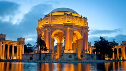 Palace of Fine Arts which is a popular location to visit during a day trip in San Francisco. The palace is illuminated from the lights around the building. It is an older building with beautiful greek architecture.