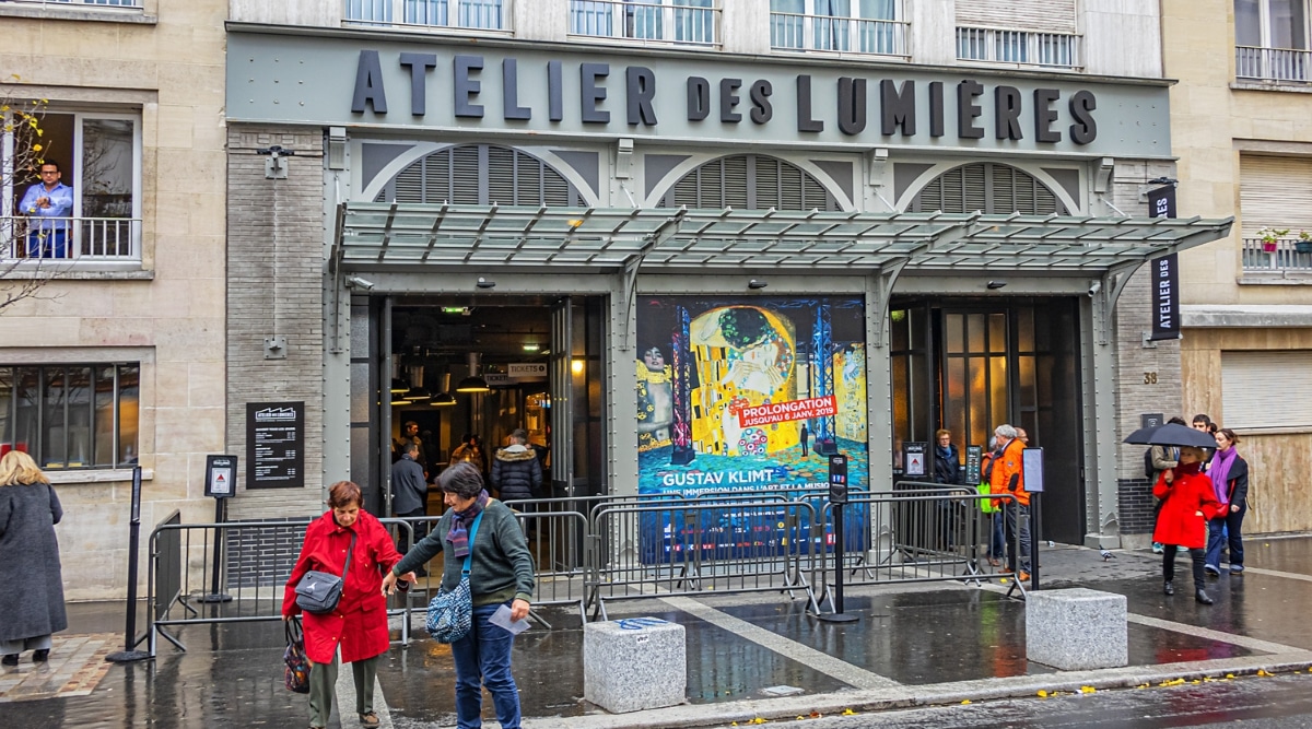 Entrance to Atelier des Lumieres in Paris France on a rainy day as tourists walk the sidewalks with umbrellas. 