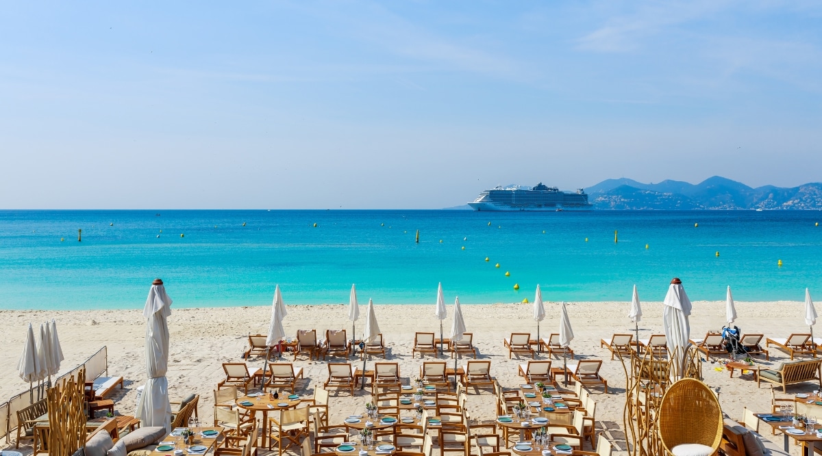 Public beach with lounge chairs and patio tables set up outside a resort in Cannes France on a sunny day with blue waters and white sands.