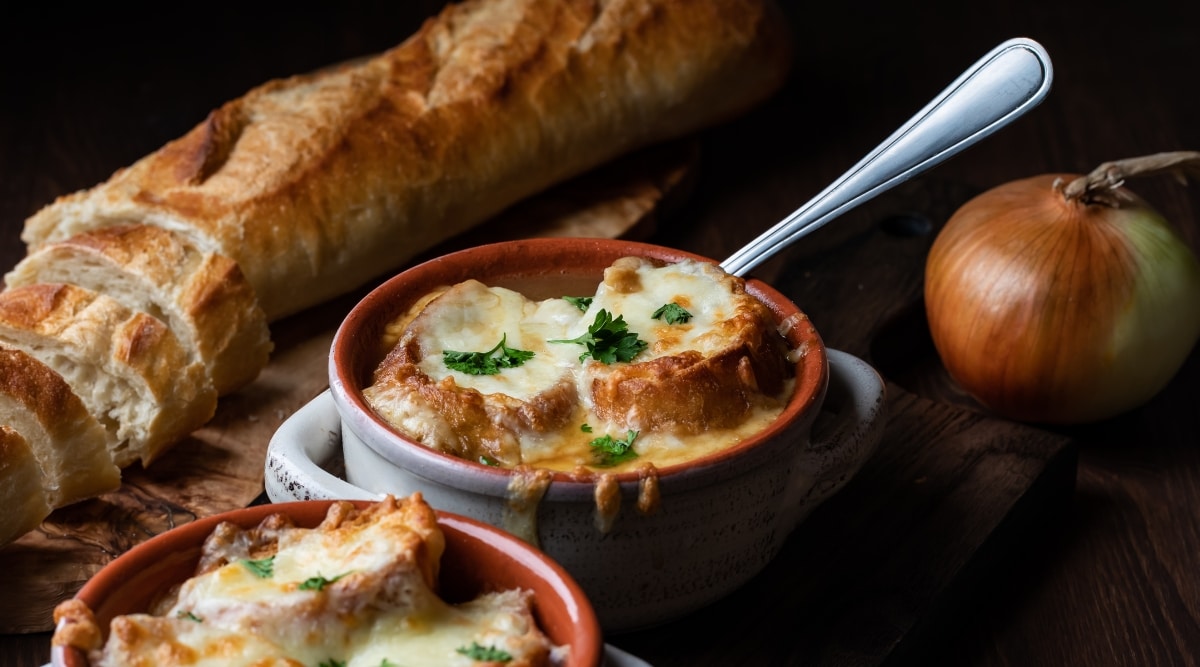 Close up of a cup of French Onion soup with a Sourdough baguette and a whole unpeeled onion in the background. 