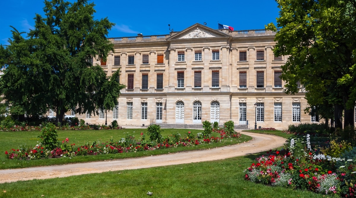 The famous Musee des Beaux-Arts in Reims France photographed from the street view on a clear sunny day with green grass and red and white flowers lining the pathway.