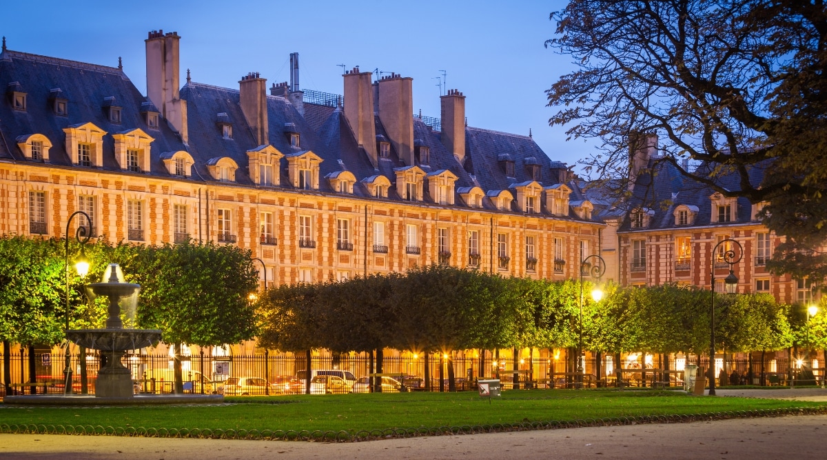 The Place des Vosges in Le Marais district in Paris, France, at dusk, lit up by streetlights and headlights from passing cars. 