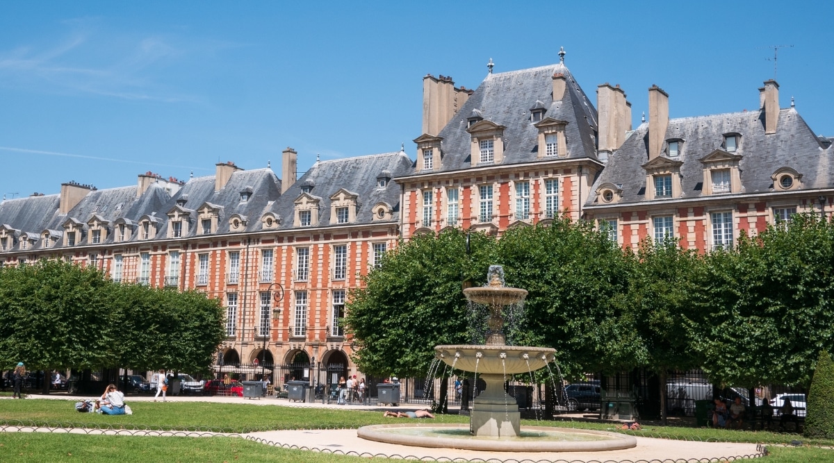 One of the four fountains in the Place des Vosges in Paris, France, with tourists on the grass and a row of houses in the background.