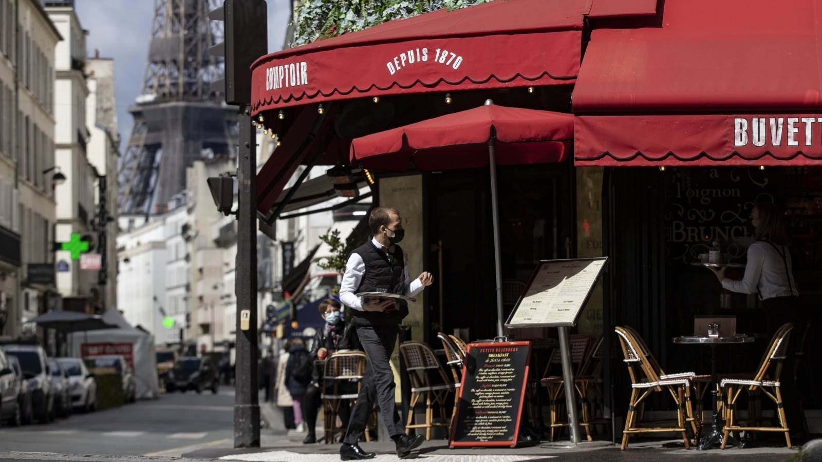 A waiter serves beverages on a cafe's terrace near the Eiffel Tower. The roof of the restaurant and the awnings on the terrace are red with white inscriptions with the name and year of opening of the restaurant. The terrace has chairs and tables for visitors, as well as a large menu board.