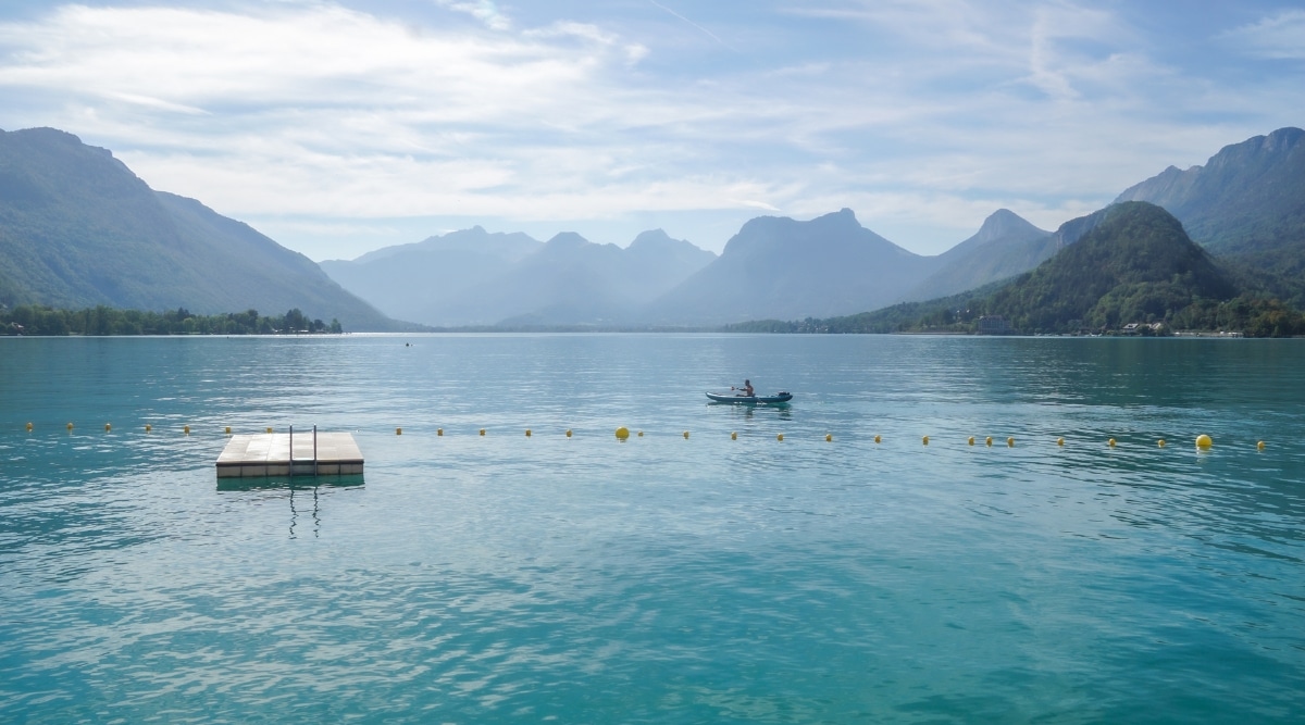 View of Lake Annecy surrounded by mountain scenery. A man is floating on the lake in a kayak. This pristine lake, nestled amidst majestic Alpine mountains, shimmers in various shades of blue and green, its crystal-clear waters reflecting the surrounding peaks and lush forests.