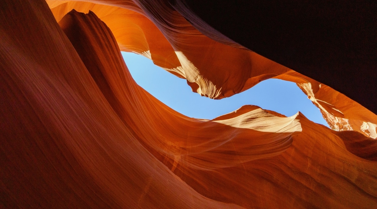 View of Antelope Canyon against a blue sky. This narrow sandstone slot canyon, sculpted by wind and water, features gracefully curving, wave-like walls in shades of red, and orange.