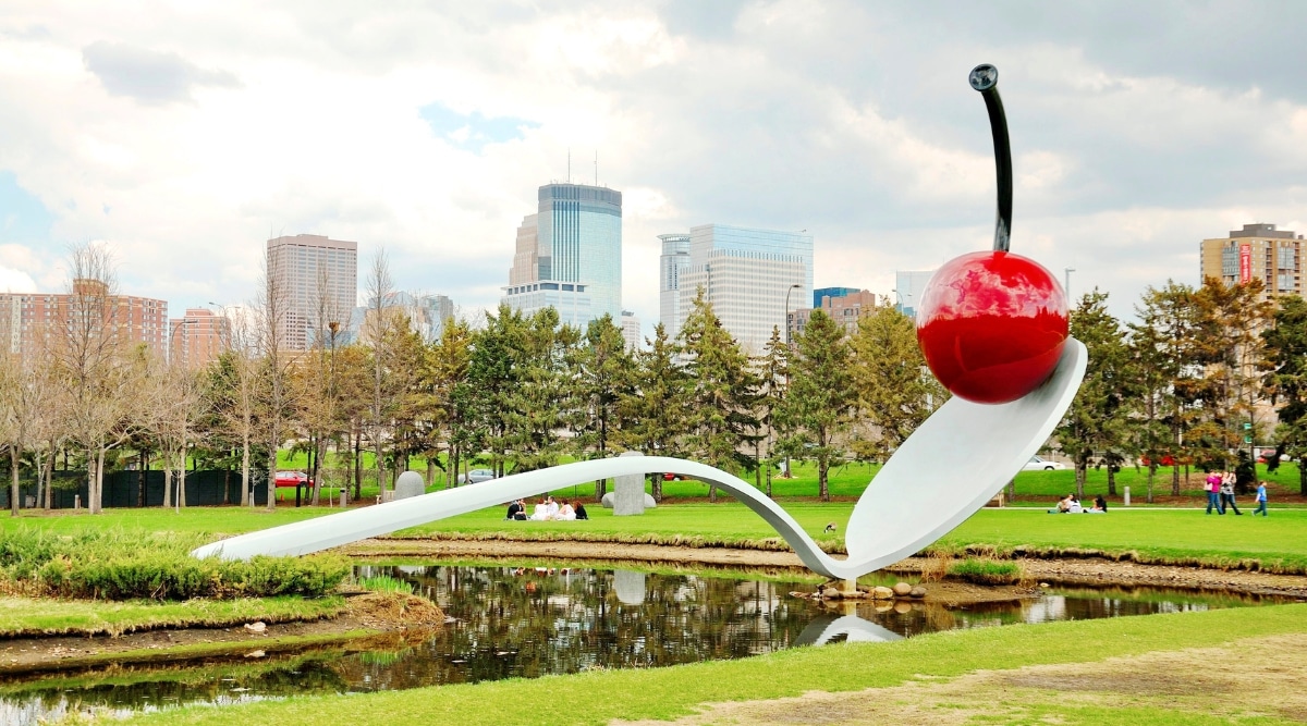 Minneapolis Sculpture Garden, located in Minneapolis, Minnesota, USA. The image reveals ponds, lush green lawns, and trees set against the backdrop of towering skyscrapers. The iconic 'Spoonbridge and Cherry' sculpture, a large spoon with a massive, reflective cherry on its bowl, takes center stage. 