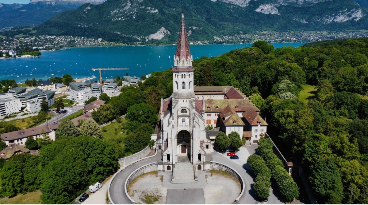 Aerial view of the Basilique de la Visitation surrounded by a lake, picturesque Alps and green lush trees. This elegant church stands out with its pristine white façade, ornate detailing, and a graceful dome that gleams in the sunlight.