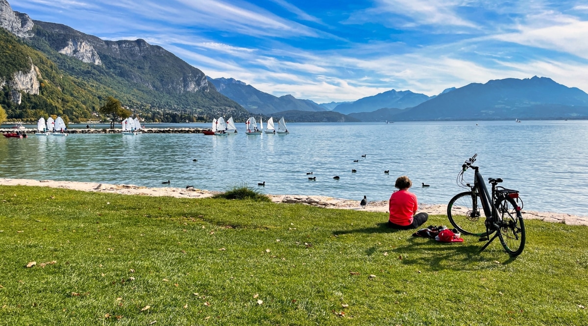 View of a cyclist sitting next to a bicycle on the shore of Lake Annecy. The cyclist is wearing a bright red sweater. The lake has crystal clear water and is surrounded by incredible Alpine peaks.