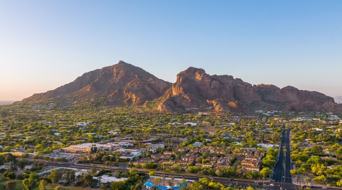 This striking image showcases the majestic Camelback Mountain in Phoenix, Arizona. Its rugged terrain and prominent red rock formations are prominently featured. In the foreground lies the charming settlement of a small town, characterized by its low-rise buildings, abundant greenery, and an abundance of bushes and trees.