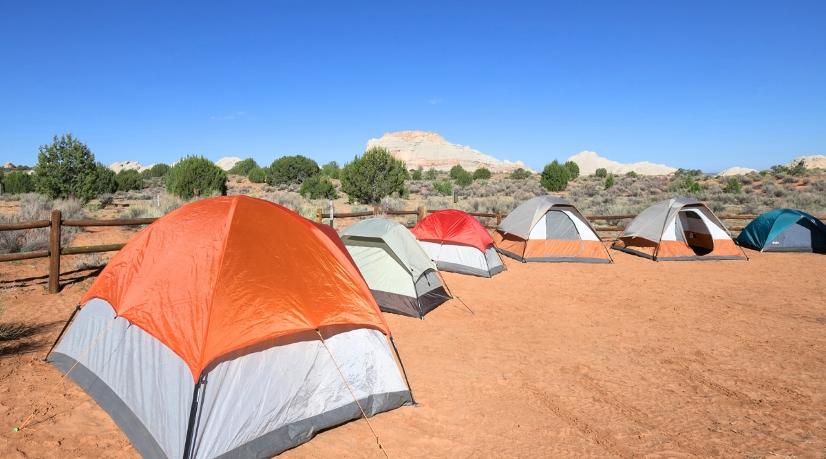 The image captures a campsite tucked against the cliffs in northern Arizona, USA. Several identical tents but in different colors including red, orange, gray and blue stand on the sandy surface. This campsite is nestled within a picturesque desert landscape, offering a truly mesmerizing setting. The sky is a vibrant, clear blue.