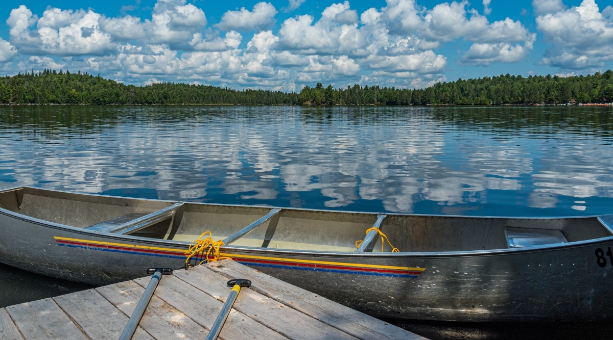 View from the pier on Canoe with the lake in the background and green trees and bushes in the distance. The blue sky with many white clouds is reflected in the crystal clear water. The canoe is metal with multi-colored thin stripes along the edges of yellow, red and blue.