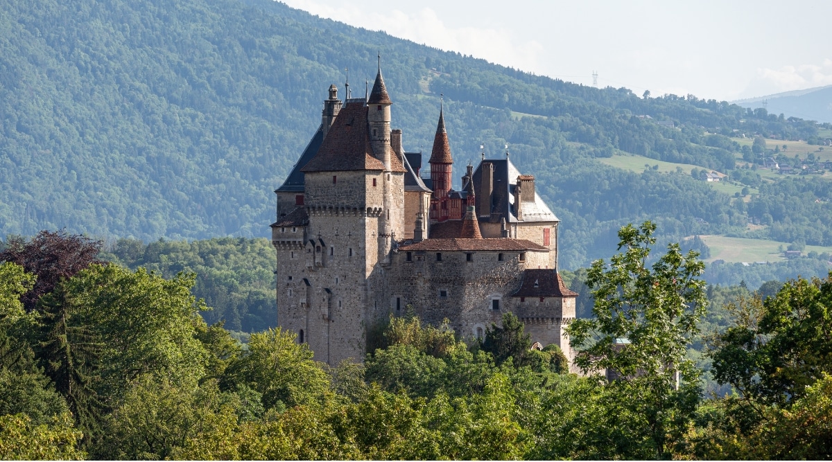 Picturesque view from a distance of the Chateau de Menthon-Saint-Bernard, a medieval castle nestled amidst lush trees. The castle boasts fairy-tale architecture with its brown towers contrasting beautifully with the Alpine backdrop.