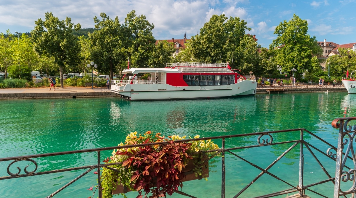 View of a small red and white cruise ship on Lake Annecy. The water is crystal clear with an emerald tint. The embankment has a lot of greenery and trees. People walk along the embankment.