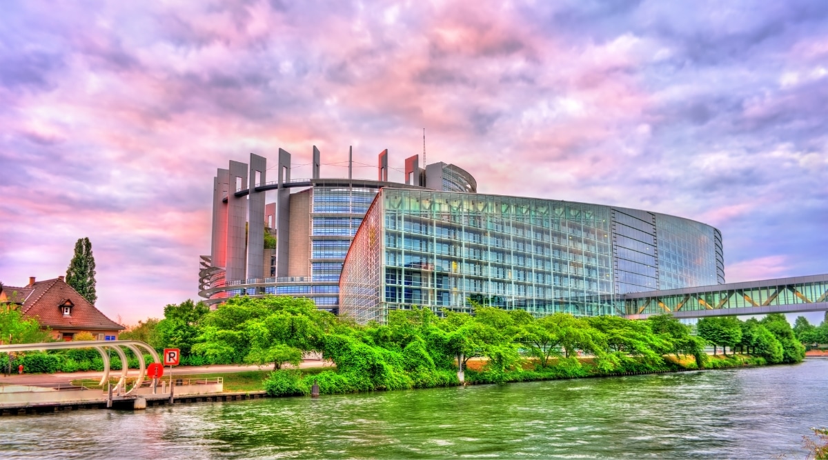 The giant glass Parliament building in Strasbourg, France, sitting along the Ill River with a cloudy pink sky and lush greenery along the river. 