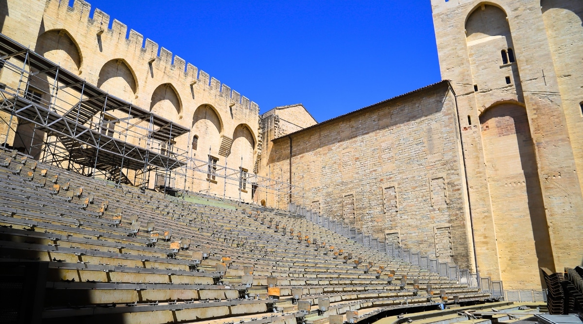View of the grand courtyard of the Palais des Papes, where the Festival d'Avignon is typically held. This site is an imposing structure with its massive stone walls, soaring towers, and Gothic architectural details. Its grand courtyard becomes an open-air theater where various performances take place.