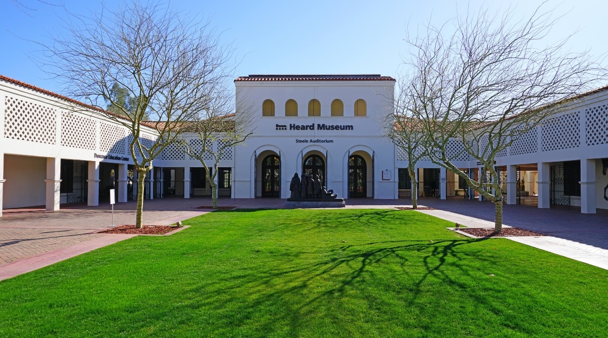View of the Heard Museum in Phoenix and the front yard. The Heard Museum in Phoenix boasts an eye-catching building that features warm, earthy tones, and desert-inspired textures. The front yard has a manicured lawn and several trees with bare branches. There is a sculpture in front of the building's entrance.