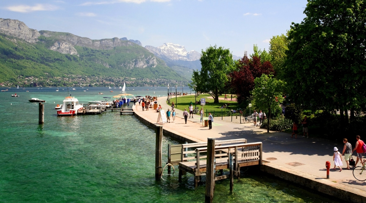 View of the Jardins de l'Europe, the embankment and Lake Annecy. The Jardins de l’Europe is a picturesque and meticulously manicured garden in the heart of Annecy, France. The park has meticulously trimmed shrubs and a variety of stately trees. The water in the lake shimmers with emerald and blue shades. People walk along the embankment enjoying the magnificent mountain scenery.