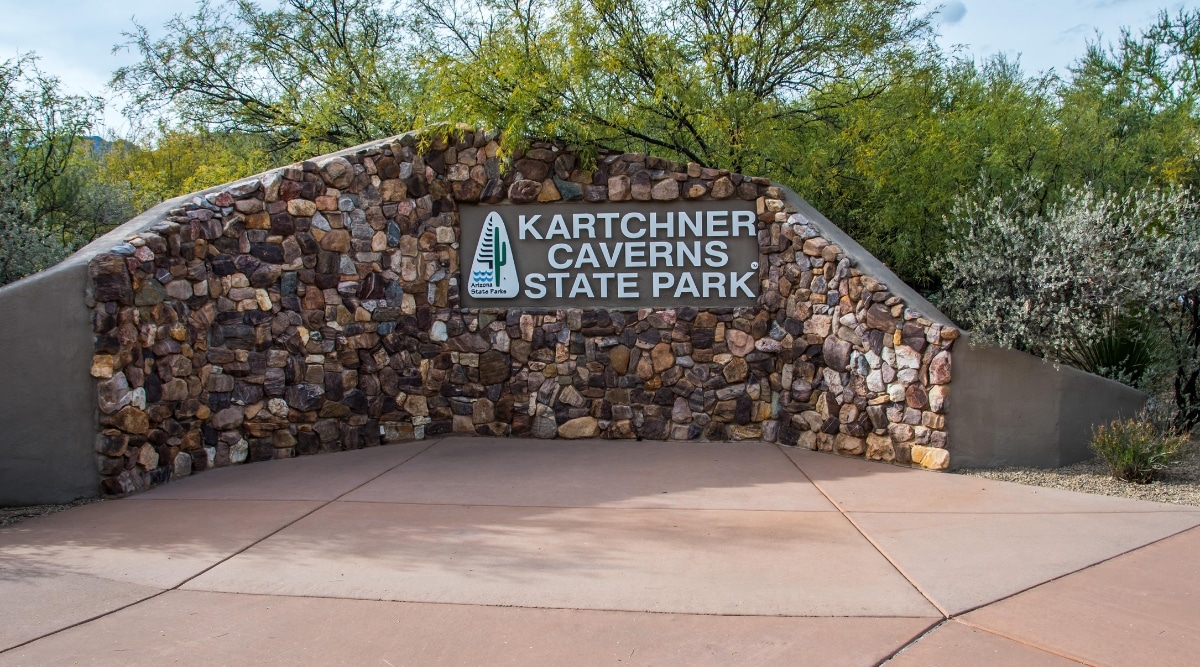 At the entrance to Kartchner Caverns State Park, there is a wall crafted from vibrant, colorful stones, adorned with the park's name "Kartchner Caverns State Park." A multitude of trees provides a lush backdrop to this impressive wall.