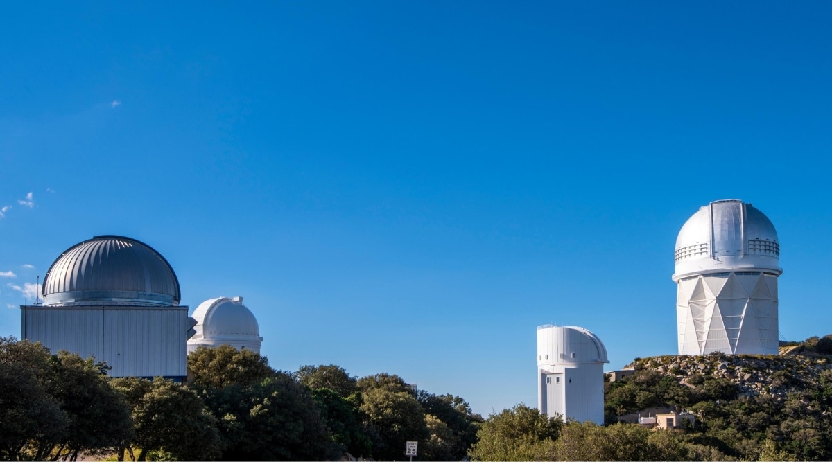A breathtaking view of the Kitt Peak National Observatory. The image captures the array of state-of-the-art telescopes and astronomical equipment under a clear sky. The dome-shaped telescopes of the observatory stand on a mountainous hill among green nature.