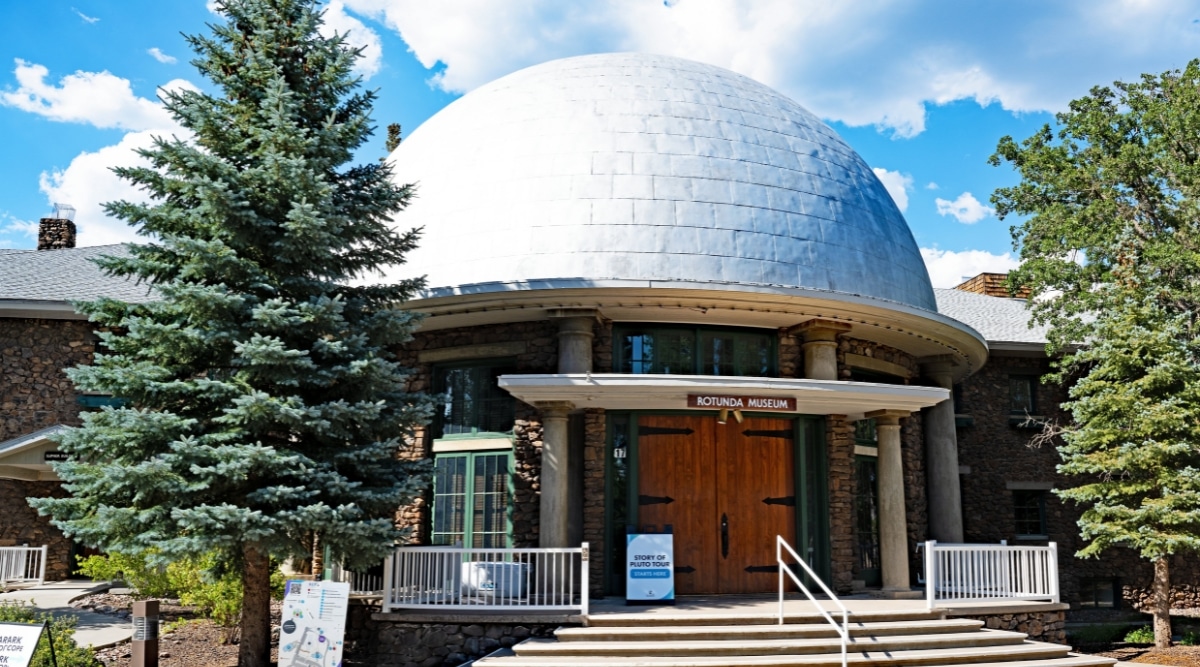 The Lowell Observatory in Flagstaff is a picturesque astronomical. The observatory stands as a beacon of scientific discovery, with its distinctive white-domed telescopes and red-brick buildings set against. A number of pine trees stand in front of the building.