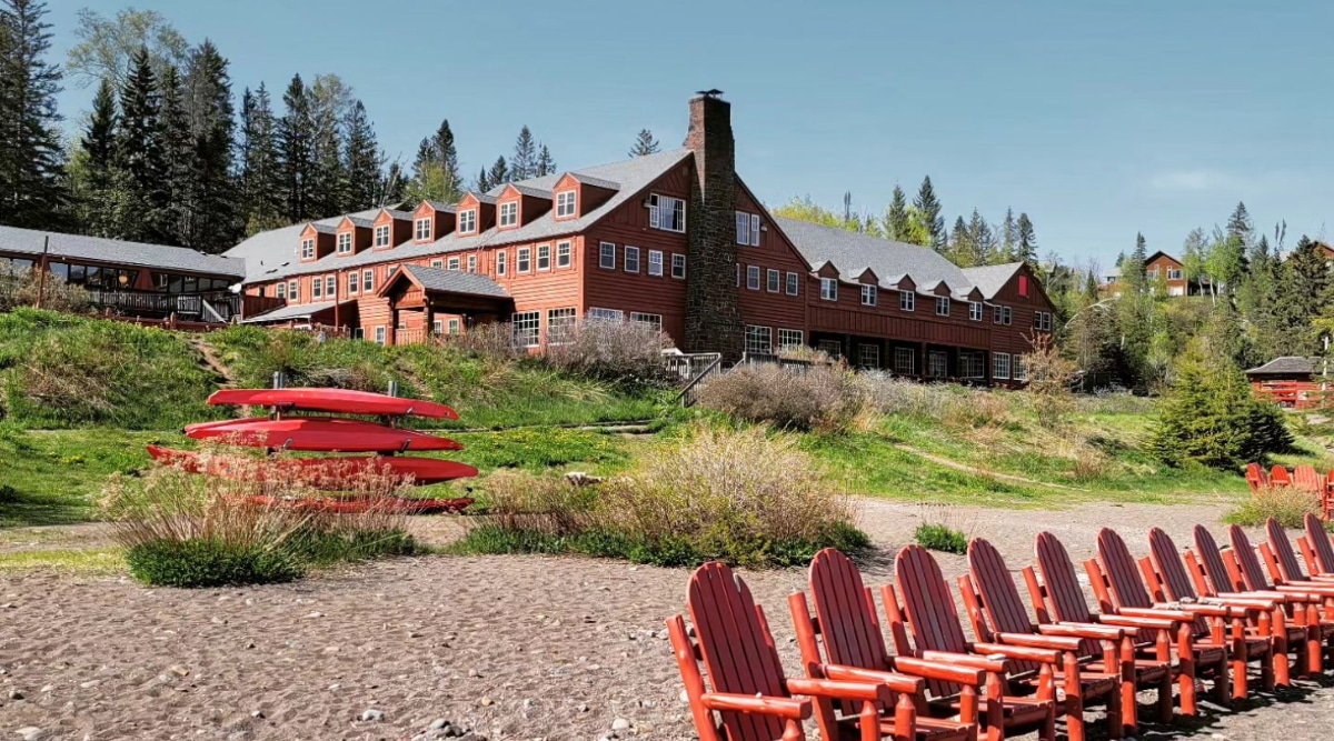 View of Lutsen Resort. A large three-story building with red brick trim and gray slanted roofs. The building also has an extended terrace overlooking the beach. In the foreground there is a sandy beach with red wooden lounge chairs and red kayaks.