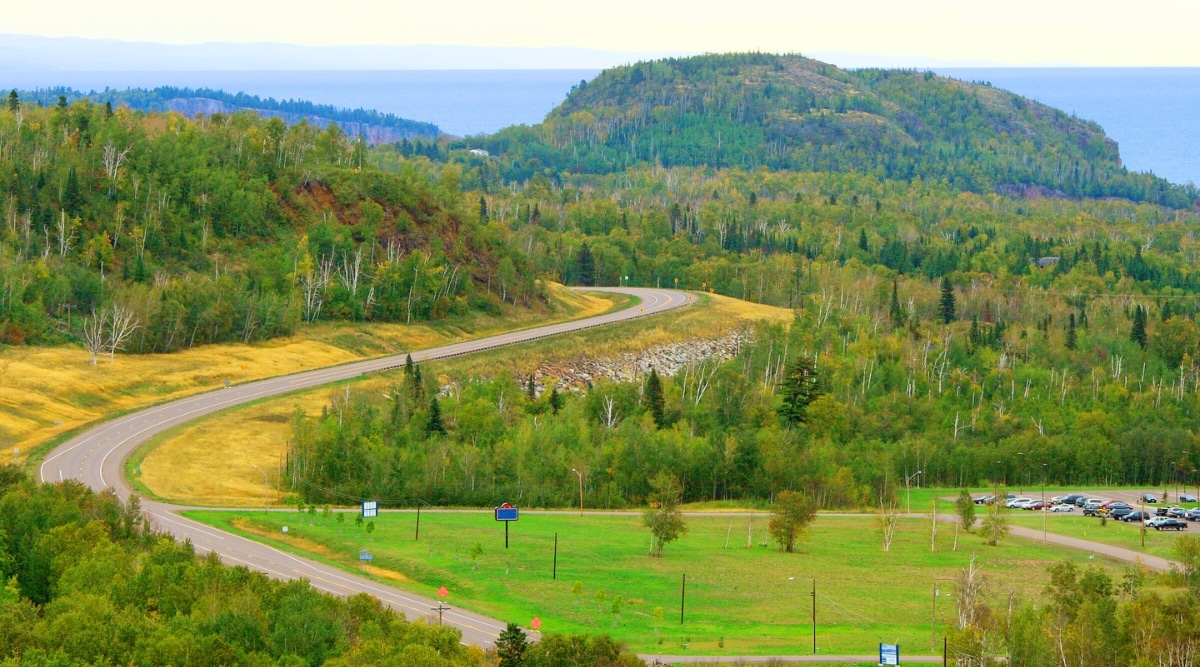 An image captures the beauty of the North Shore Scenic Drive, a winding coastal road that meanders along the shoreline of Lake Superior in the northern United States. The road stretches ahead, hugging the rocky coastline. On the right side of the road, a lush and dense forest of evergreen trees.