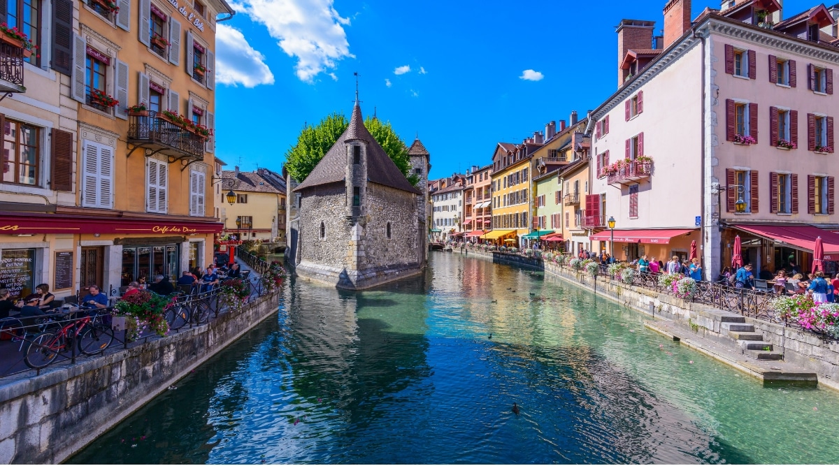 View of the Palais de I'lle on a sunny day. The Palais de I'lle, a beautifully preserved medieval building in Annecy, stands gracefully in the middle of the Thiou River. Its distinctive triangular shape, narrow windows, and stone turret evoke a sense of historical charm and architectural elegance.