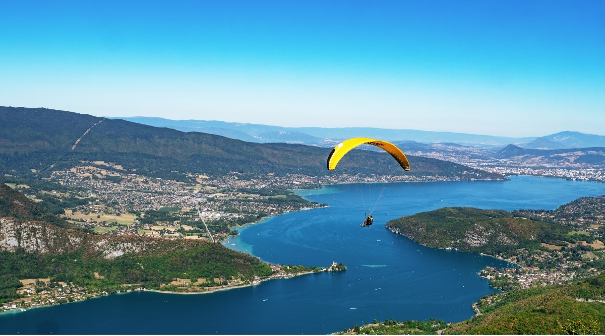 View of paraglide over Lake Annecy. The aerial view of Lake Annecy is breathtaking, offering a panoramic vista of its stunning natural beauty. Nestled within the embrace of the majestic French Alps, the lake's crystal-clear waters shimmer in various shades of blue and green. The surrounding landscape showcases quaint lakeside villages, lush forests, and the charming town of Annecy.