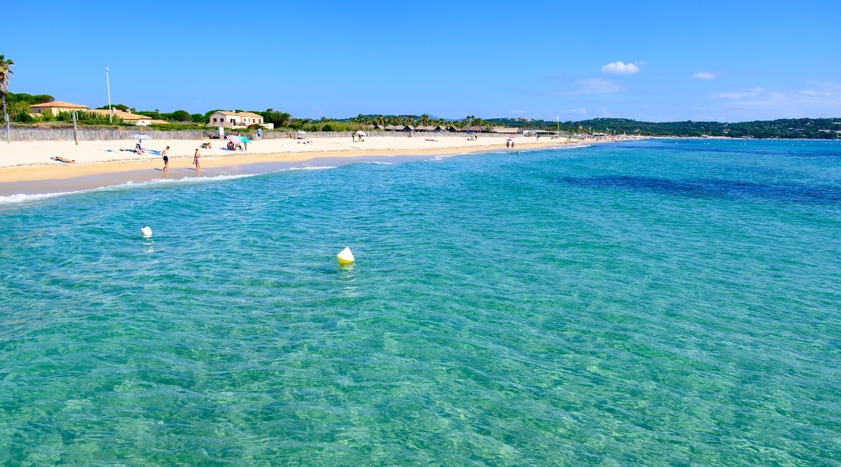 This image showcases Plage de Pampelonne on a radiant, sunlit day, where the crystal-blue sea glistens with gentle waves. The beach is bustling with people enjoying the sunshine. In the background, several houses and lush greenery provide a charming backdrop to the vibrant scene.