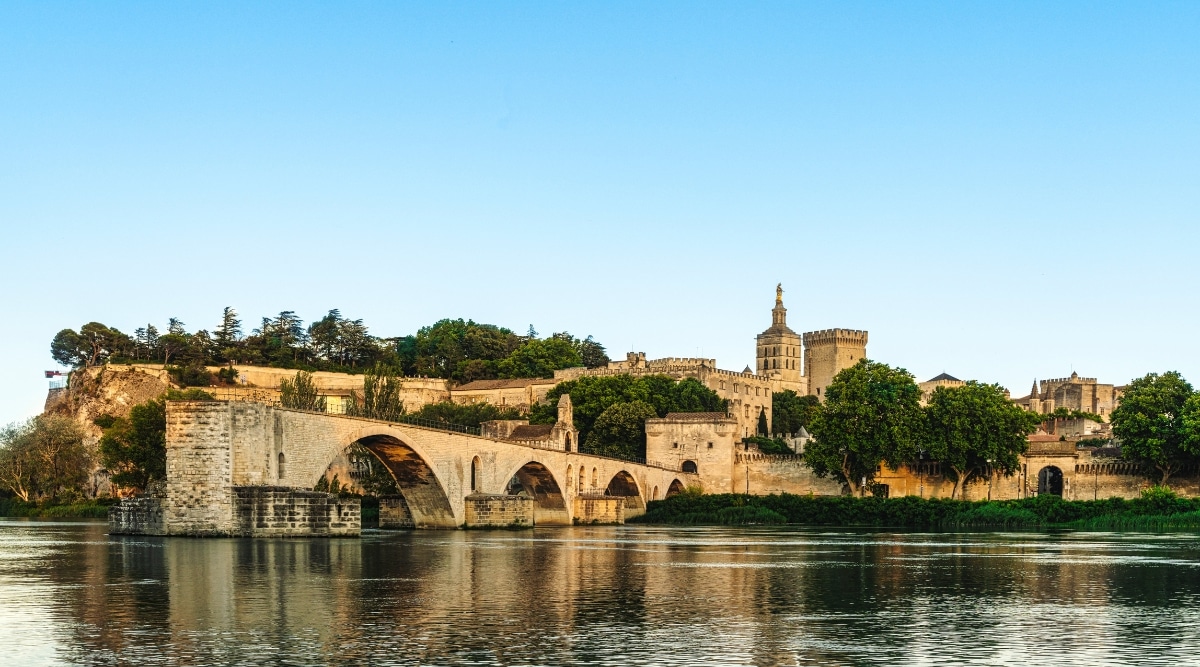 View of Pont Saint-Benezet. The Pont Saint-Benezet, also known as the Pont d'Avignon, is a picturesque and iconic bridge in Avignon, France. This historic structure has a series of graceful stone arches that extend only partway across the Rhone River. Green trees and elements of a stone bridge are reflected in the river.